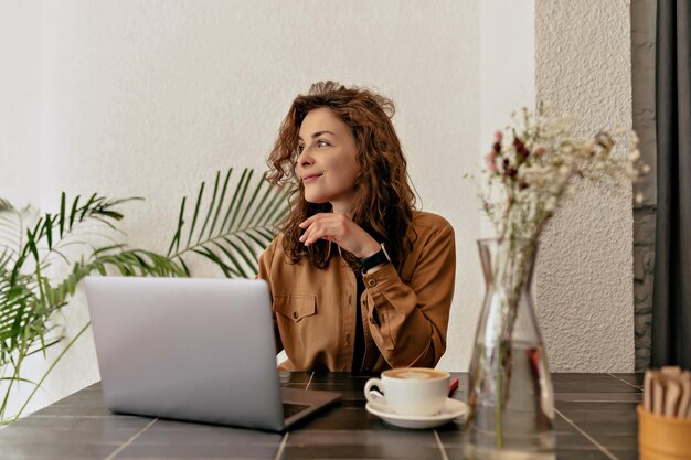 Fröhliche junge Frau, die beiseite schaut, sitzt auf dem Tisch mit Laptop und lächelt Brünette mit lockigem Haar trägt lässige Kleidung Positive Emotionen Konzept