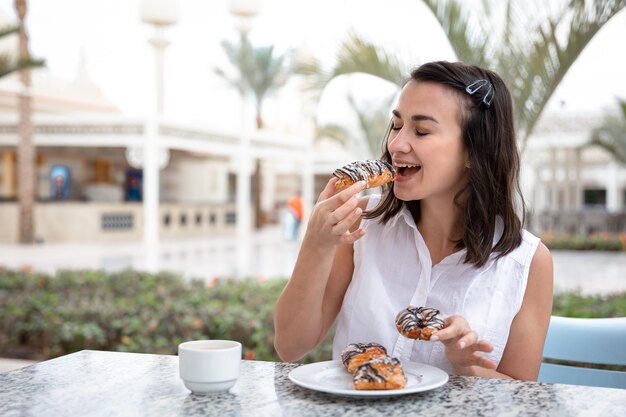 Fröhliche junge Frau, die Morgenkaffee mit Donuts auf der Außenterrasse genießt.