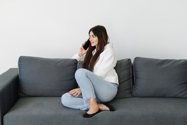 Fröhliche junge Frau, die zu Hause auf einer Couch sitzt und mit dem Handy telefoniert