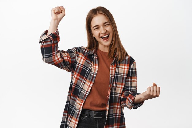 Fröhliche junge Frau erreicht Zielerfolge, schreit vor Freude, Faustpumpe, wurzelt für das Team, gewinnt Leistung, Preis, gewinnt und triumphiert, weiße Wand
