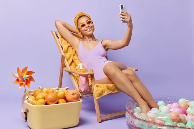 Fröhliche junge Frau fühlt sich entspannt, trägt Sonnenbrille und Badeanzug macht Selfie über Smartphone-Posen auf Liegestuhl vor violettem Hintergrund Fröhliches weibliches Model nimmt Video für ihren Blog am Strand auf