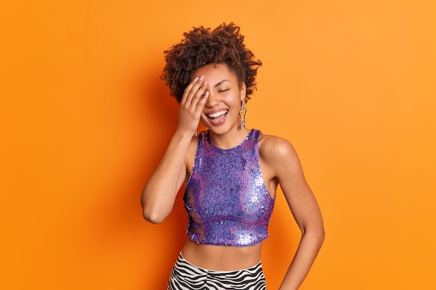 Fröhliche junge Frau hat Afro-Haar lächelt breit hält Hand auf Gesicht fühlt sich freudig kichern über lustige Geschichte trägt lila Top sternförmige Ohrringe isoliert über orange Wand