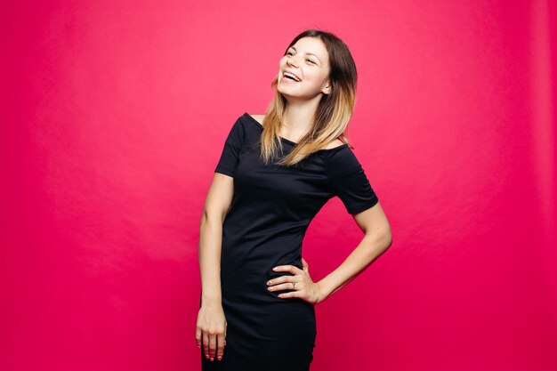 Fröhliche junge Frau im schwarzen Kleid, die im Studio posiert und lauthing wegschaut Positivität emotionales Mädchen, das die Hand an der Taille hält, die vor rosa Hintergrund steht Konzept des Einkaufens fashionbeauty