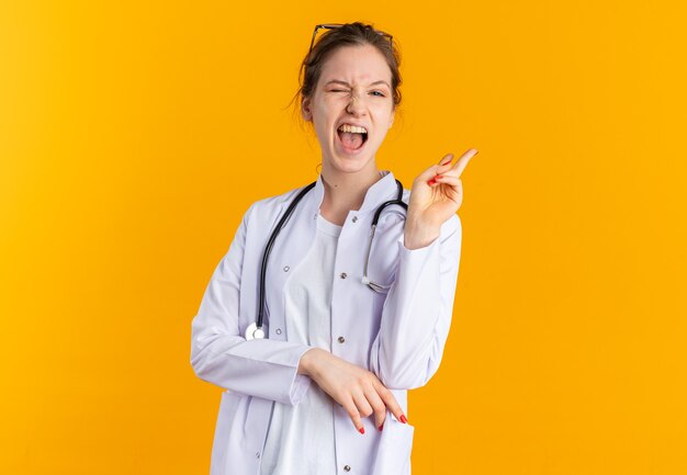 Fröhliche junge Frau in Arztuniform mit Stethoskop blinzelt mit den Augen und gestikuliert Siegeszeichen