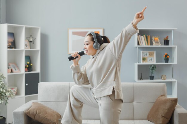 Fröhliche junge Frau in Freizeitkleidung mit Kopfhörern, die auf einer Couch zu Hause tanzen und die Fernbedienung halten, indem sie sie als Mikrofon singen und Spaß haben