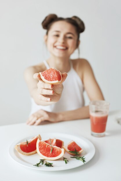 Fröhliche junge Frau lächelnd sitzen am Tisch, der Stück Grapefruit über weiße Wand streckt. Gesundes Lebensmittelkonzept.