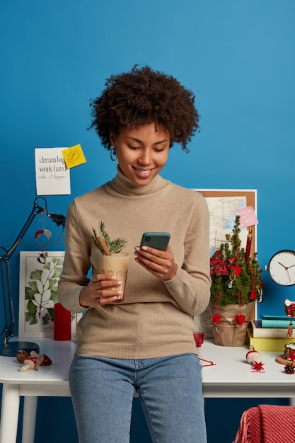 Fröhliche junge Frau mit Afro-Haar überprüft Newsfeed auf dem Smartphone, zufrieden, Nachrichten und Kommentare der Follower unter ihrem Beitrag zu lesen, trinkt Eierlikör-Cocktail steht in der Nähe des Arbeitsplatzes konzentriert auf dem Bildschirm