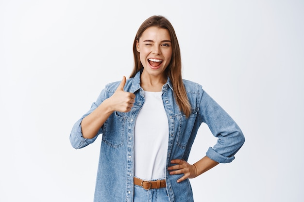 Fröhliche junge Frau mit blonden Haaren und legerer Kleidung, Daumen hoch zur Zustimmung, wie eine gute Sache, genehmigen und empfehlen, gute Arbeit loben oder ja sagen, weiße Wand