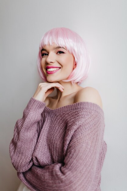 Fröhliche junge Frau mit glänzendem hellrosa Haar, das auf heller Wand lächelt. Innenporträt des herrlichen weiblichen Modells im niedlichen peruke und im lila Pullover.