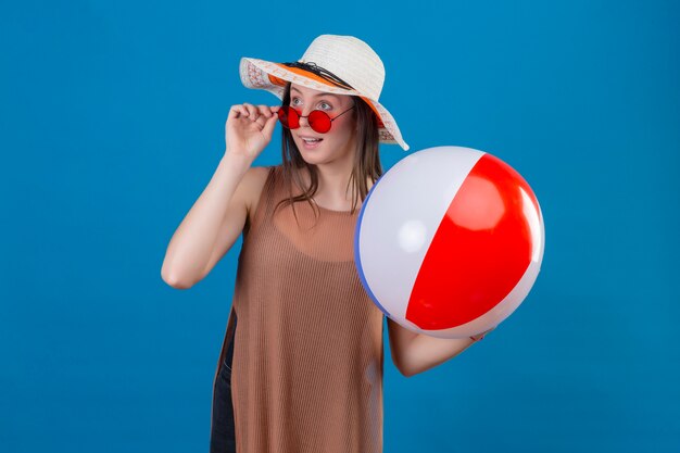 Fröhliche junge Frau mit Hut, die rote Sonnenbrille hält, die aufblasbaren Ball hält, der beiseite mit glücklichem Lächeln steht, das auf Blau steht