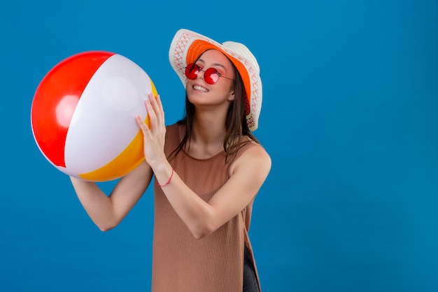 Fröhliche junge Frau mit Hut, die rote Sonnenbrille hält, die aufblasbaren Ball lächelnd mit glücklichem Gesicht steht auf Blau steht