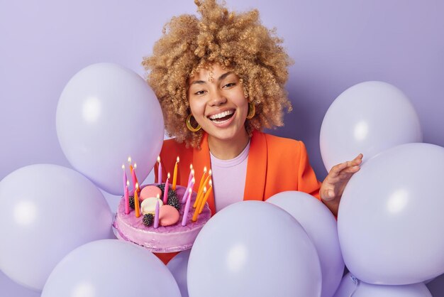 Fröhliche junge Frau mit lockigem Haar in stylischen orangefarbenen Jackenposen mit köstlichem Geburtstagskuchen feiert besondere Anlässe um aufgeblasene Luftballons isoliert über violettem Hintergrund