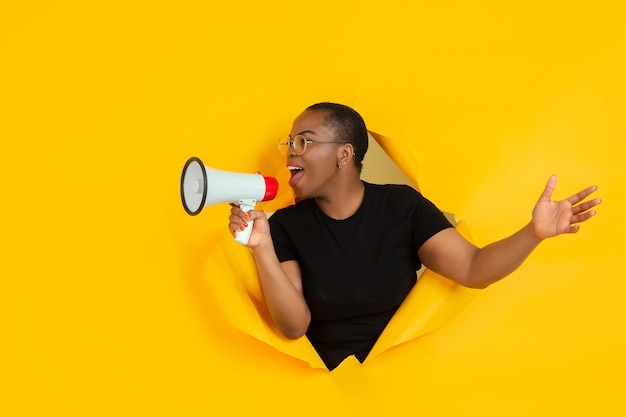 Fröhliche junge Frau posiert in zerrissener gelber Papierlochwand emotional und ausdrucksstark schreien und mit dem Lautsprecher rufen