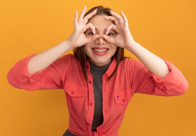 Fröhliche junge hübsche Frau, die eine Blickgeste macht, die mit den Händen als Fernglas auf die orangefarbene Wand schaut