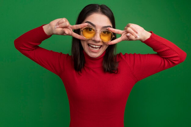 Fröhliche junge hübsche kaukasische Frau mit Sonnenbrille, die ein V-Zeichen-Symbol in der Nähe der Augen zeigt