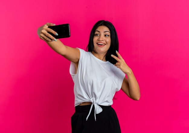 Fröhliche junge kaukasische Frau gestikuliert Siegeshandzeichen unter selfie