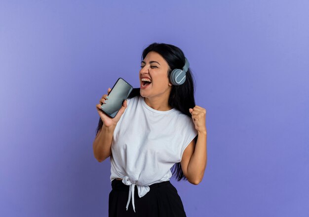 Fröhliche junge kaukasische Frau mit Kopfhörern hält das Telefon, das vorgibt zu singen
