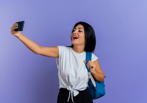 Fröhliche junge kaukasische Frau mit Rucksack schaut auf das Telefon und macht Selfie