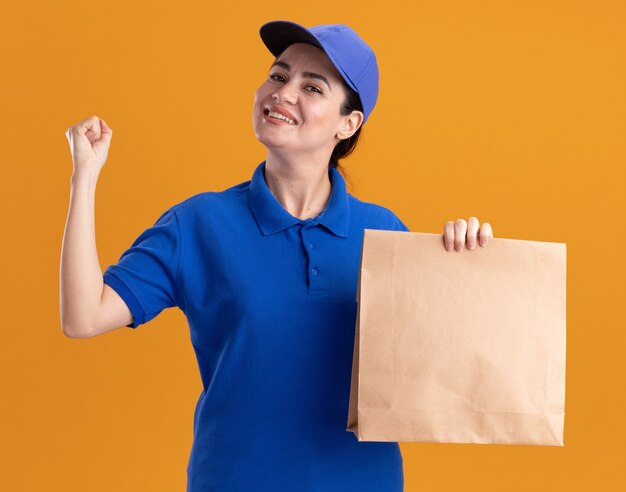 Fröhliche junge Lieferfrau in Uniform und Mütze, die ein Papierpaket mit Blick auf die Vorderseite hält und eine starke Geste isoliert auf der orangefarbenen Wand macht