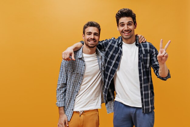 Fröhliche junge Männer in karierten blauen Hemden, weißen T-Shirts und bunten Hosen posieren gut gelaunt und lächelnd an oranger Wand.
