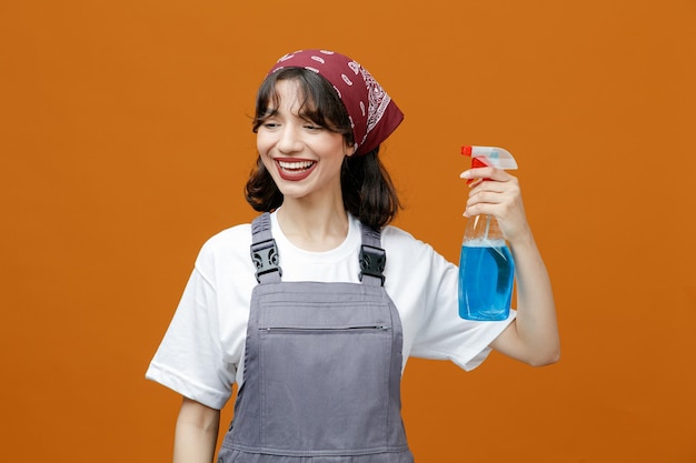 Fröhliche junge Putzfrau in Uniform und Bandana mit Reinigungsmittel, die es auf sich selbst anwendet und die Seite isoliert auf orangefarbenem Hintergrund betrachtet