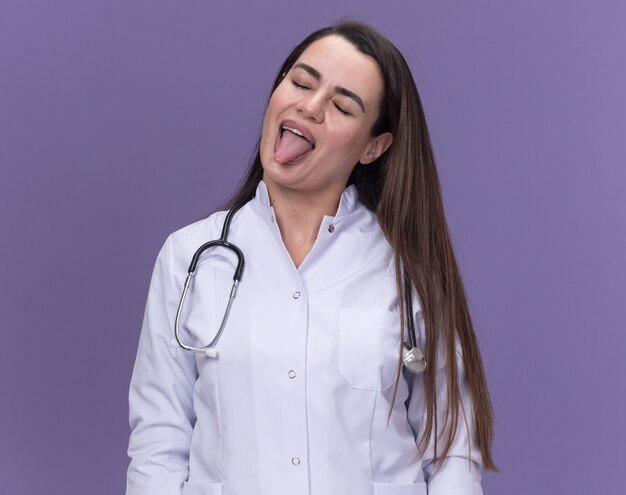 Fröhliche junge Ärztin, die ein medizinisches Gewand mit Stethoskop trägt, streckt die Zunge heraus