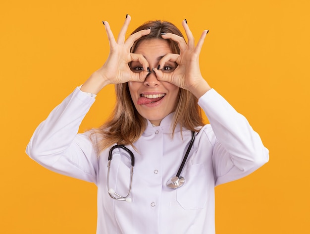 Fröhliche junge Ärztin, die medizinische Robe mit Stethoskop trägt, die Blickgeste und Zunge einzeln auf gelber Wand zeigt