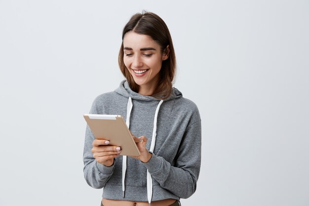 Fröhliche junge schöne brünette kaukasische Studentin mit langen dunklen Haaren im lässigen grauen Kapuzenpulli lächelnd, digitales Tablett in den Händen haltend, durch lustige Fotos von Freunden schauend