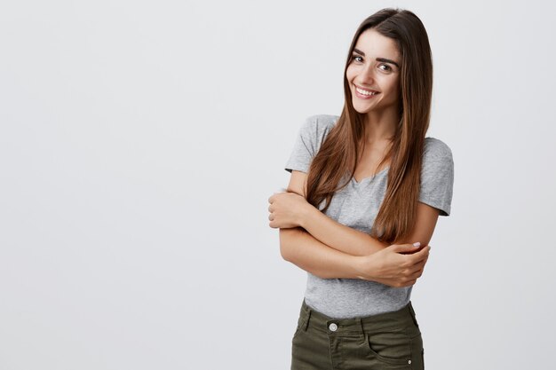 Fröhliche junge schöne brünette kaukasische Studentin mit langen Haaren in lässigem, stilvollem Outfit, das hell lächelt, Hände zusammenhält und für Universitätsabschlussfoto in der hellen Wand aufwirft.