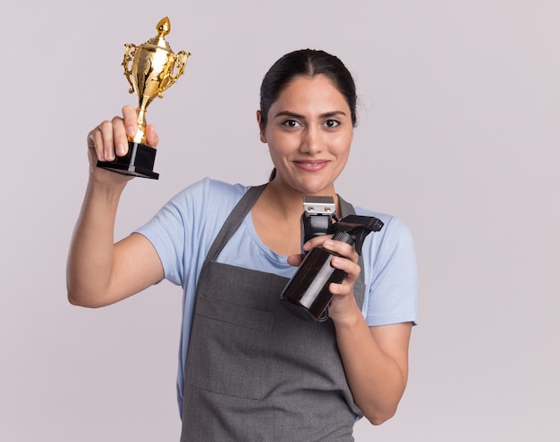 Fröhliche junge schöne Frau Friseur in der Schürze hält Gold Trophäenschneider Maschine mit Sprühflasche, die vorne mit Lächeln auf Gesicht steht, das über weißer Wand steht