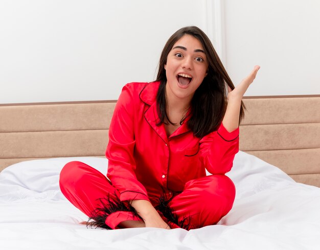 Fröhliche junge schöne Frau im roten Schlafanzug, der auf Bett sitzt und Kamera glücklich und aufgeregt im Schlafzimmerinnenraum auf hellem Hintergrund betrachtet