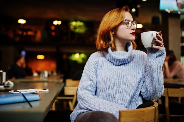 Fröhliche junge, schöne, rothaarige Frau mit Brille, die an ihrem Arbeitsplatz im Café sitzt und Kaffee trinkt