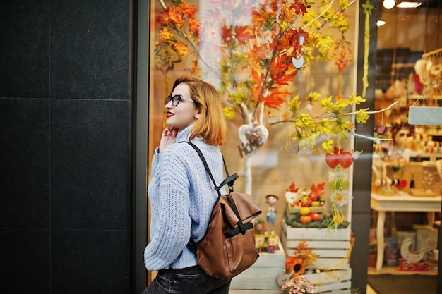 Fröhliche junge, schöne, rothaarige Frau mit Brille, warmer blauer Wollpullover mit Rucksack, posiert im Freien gegen Herbstlaub, Baumschmuck im Laden