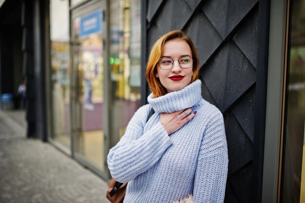 Fröhliche junge, schöne, rothaarige Frau mit Brille, warmer blauer Wollpullover mit Rucksack, posiert im Freien