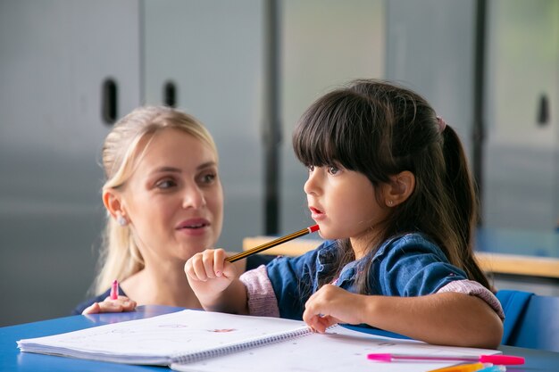 Fröhliche junge Schullehrerin, die dem kleinen Schulmädchen hilft, ihre Aufgabe zu erfüllen