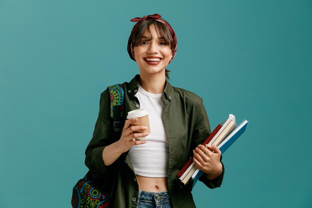 Fröhliche junge Studentin mit Bandana und Rucksack mit Notizblöcken und Kaffeetasse aus Papier, die isoliert auf blauem Hintergrund in die Kamera blickt