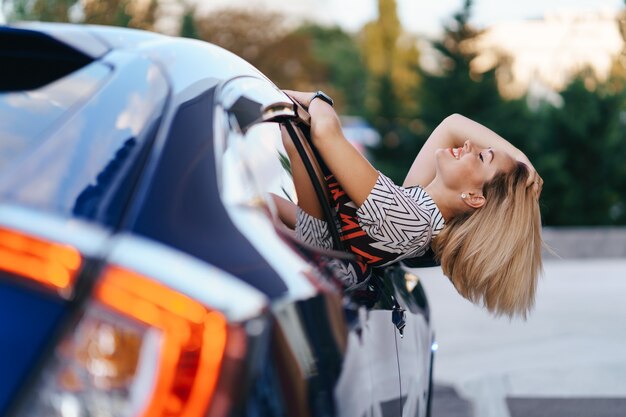 Fröhliche kaukasische Frau fährt durch die malerische sonnige Stadt und winkt mit den Armen, während sie sich an einem schönen Tag aus dem Autofenster streckt