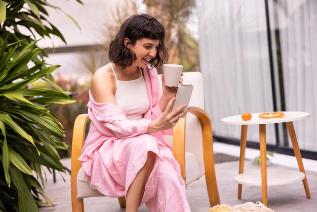 Fröhliche kaukasische junge brünette Frau, die auf den Bildschirm des Telefons schaut und den Morgenkaffee genießt Sommer-Lifestyle-Konzept