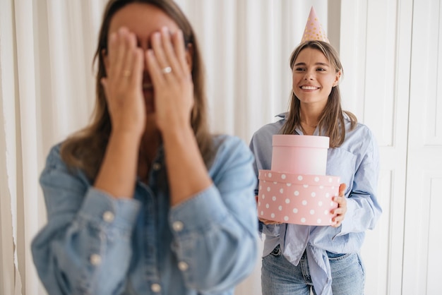 Fröhliche kaukasische junge Frau überreicht ihrer Schwester, die auf eine Schachtel wartet, ein Geschenk und bedeckt die Augen mit den Händen. Überraschungskonzept