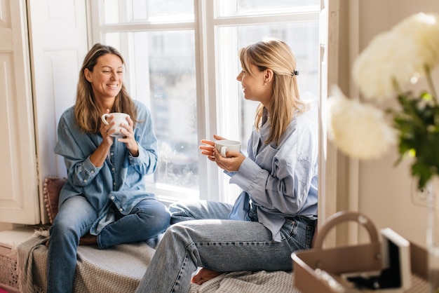 Fröhliche kaukasische schöne Frauen in Freizeitkleidung klatschen und trinken Tee, während sie tagsüber auf der Fensterbank sitzen. Freizeit, Lifestyle-Konzept