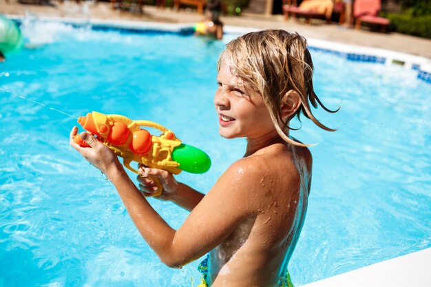 Fröhliche Kinder spielen Wassergewehre, freuen sich, springen, schwimmen im Pool.