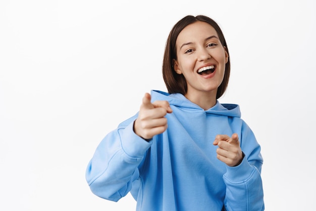 Fröhliche lachende Frau, die mit dem Finger auf die Kamera zeigt und lächelt, dich auswählt, Person gratuliert, lobt und auswählt, im Hoodie vor weißem Hintergrund steht