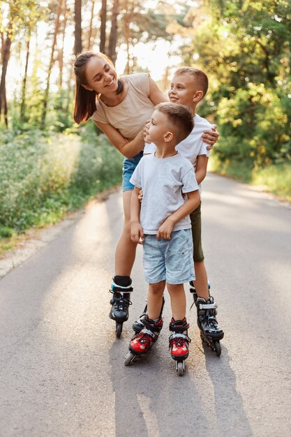 Fröhliche lächelnde Frau, die mit ihren Kindern im Sommerpark rollt, Mutter, die Kinder mit zahnigem Lächeln ansieht, Familien-Rollerblading und Spaß zusammen hat.