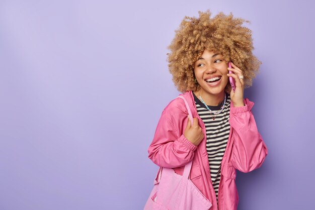 Fröhliche lächelnde junge Frau spricht auf dem Handy und lacht über den Anruf. Sie trägt eine rosafarbene Jacke und trägt eine Stofftasche