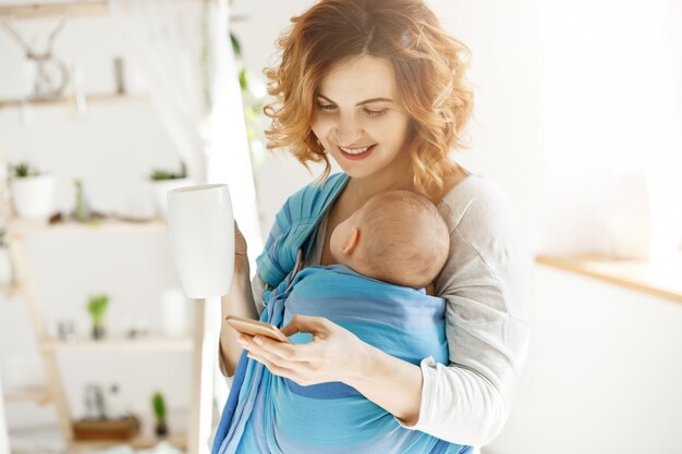 Fröhliche lächelnde Mutter trinkt Kakao und schreibt ihrem geliebten Ehemann eine SMS, während kleiner Sohn in Baby schlank döst. Atmosphäre der Liebe und des Schutzes.