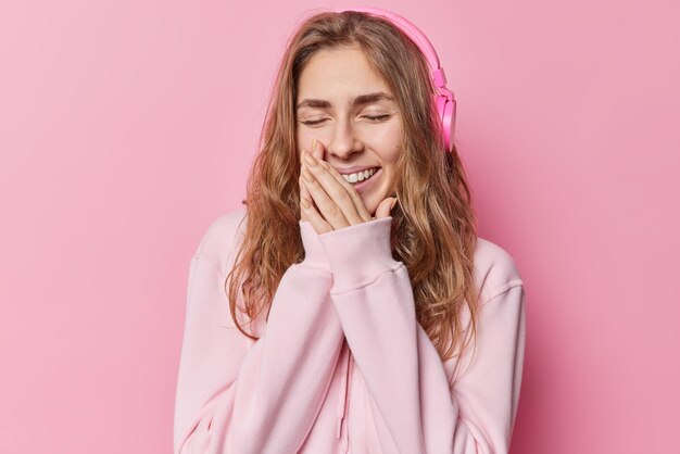 Fröhliche, langhaarige junge Frau kichert positiv, hält die Hände in der Nähe des Mundes, hört Lieblingsmusik über Kopfhörer, die in einem bequemen Sweatshirt gekleidet sind, hält die Augen isoliert vor rosa Hintergrund geschlossen