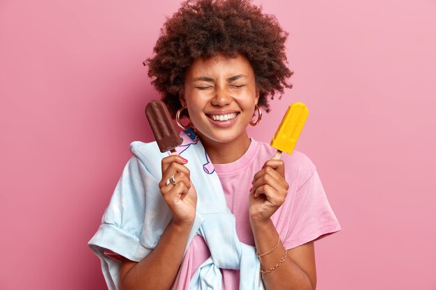 Fröhliche, lockige Afro-Amerikanerin grinst vor Glück und hält die Augen geschlossen, Posen mit zwei köstlichen Eiscremes auf Stöcken drückt positive Emotionen einzeln über rosafarbener Wand aus. Schöne Sommerzeit