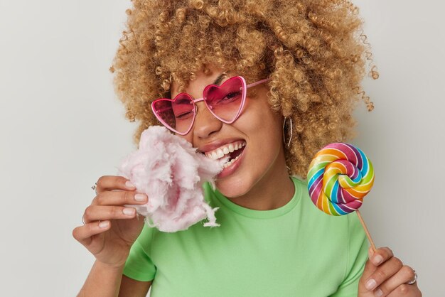 Fröhliche, lockige Frau beißt köstliche Zuckerwatte mit Appetit hält bunte Süßigkeiten am Stiel und isst gerne appetitliche Leckereien trägt herzförmige Sonnenbrille grünes T-Shirt Sommerlebensstil