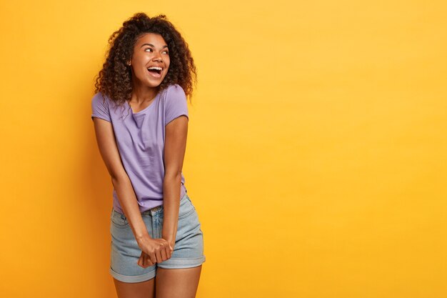 Fröhliche lockige Frau hält die Hände zusammen, lacht glücklich, konzentriert beiseite, trägt lila T-Shirt und Jeansshorts, fühlt sich glücklich und sorglos