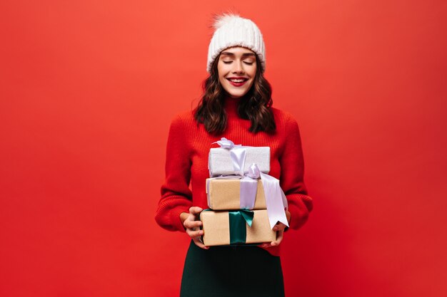 Fröhliche lockige Frau in leuchtend rotem Pullover, Strickmütze posiert mit geschlossenen Augen und hält Geschenkboxen an isolierter roter Wand